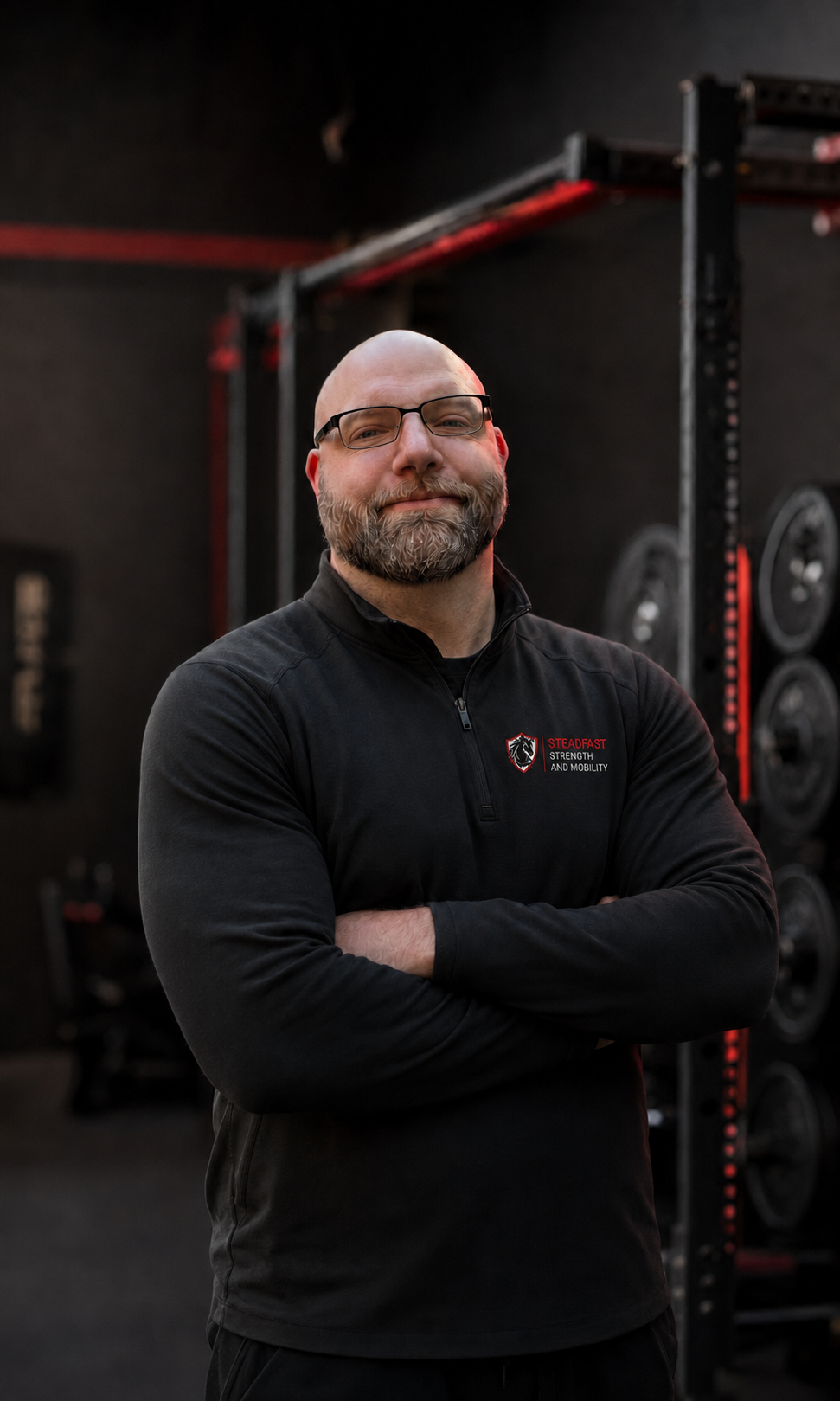 Justin Stead stands with his arms crossed inside a private strength studio wearing a black Steadfast Strength and Mobility jacket.
