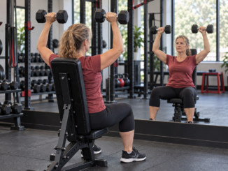 Woman performing seated dumbbell shoulder presses while watching her reflection in a gym mirror.